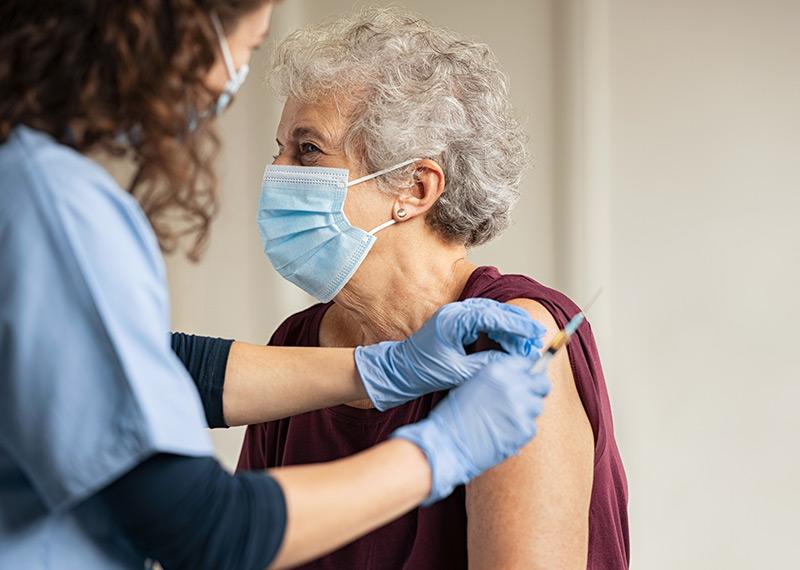 A woman wearing latex gloves prepares to administer a needle to an elderly woman wearing a mask. 