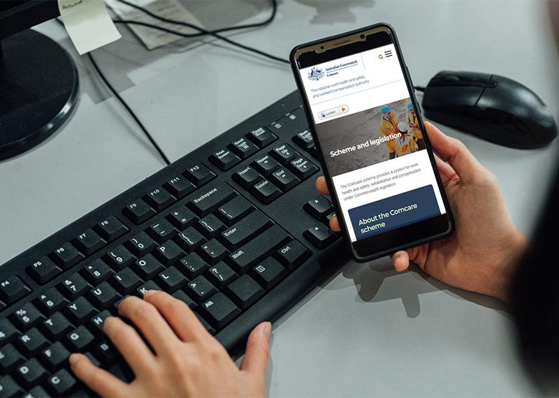 A person types on a keyboard with one hand. The other hand holds a mobile phone that says Scheme and Legislation, and About the Comcare scheme. 