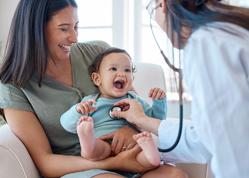 Person holding a smiling baby, who is being examined by a medical professional